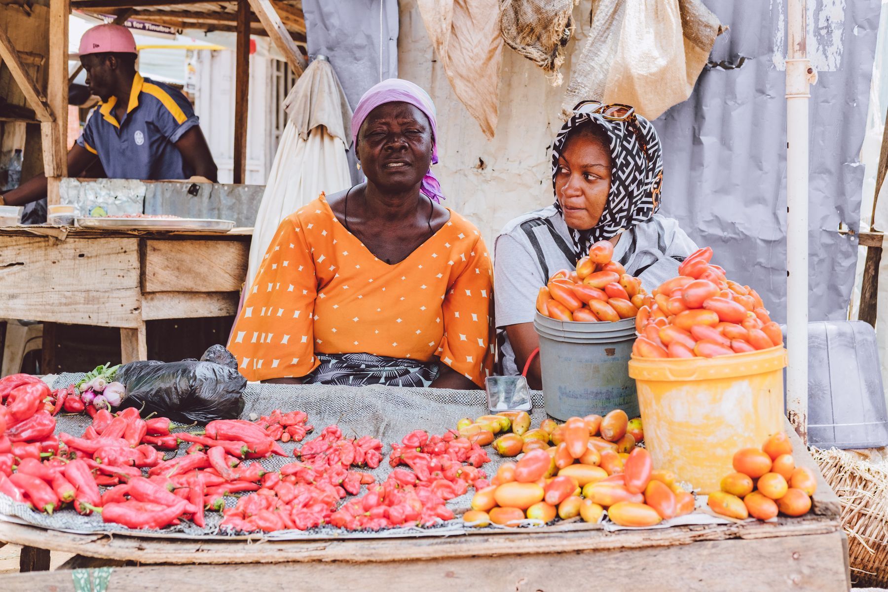 Two women smiling behind a market stall of fresh tomatoes and peppers